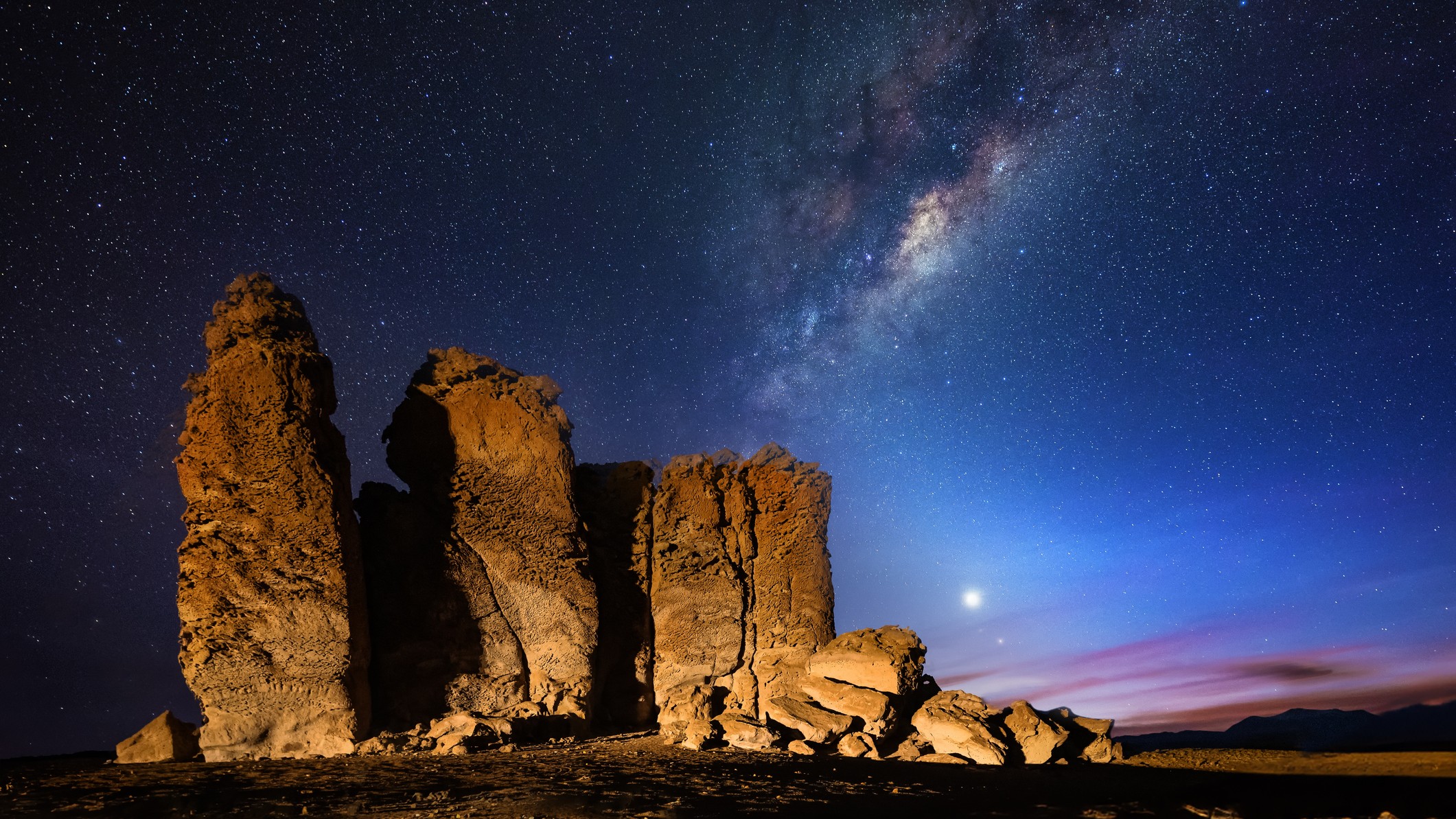 large rock formations on the left behind is the milky way filled with stars and low in the sky is a bright looking star - this is venus.