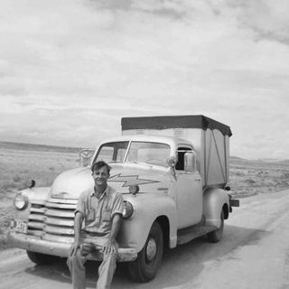 Black and white image of the artist H.C. Westermann, sat smiling on the bumper of an old vintage style truck, dirt style road, vast grass terrain landscape in the backdrop, cloudy sky