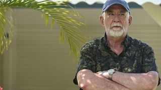 Roy Palmer (Gerard Horan), wearing a short-sleeved shirt with a dark green leaf-print pattern on it and a blue baseball cap, stands outside. He has his arms folded and looks very serious. There is a palm frond occupying most of the left-hand side of the image.