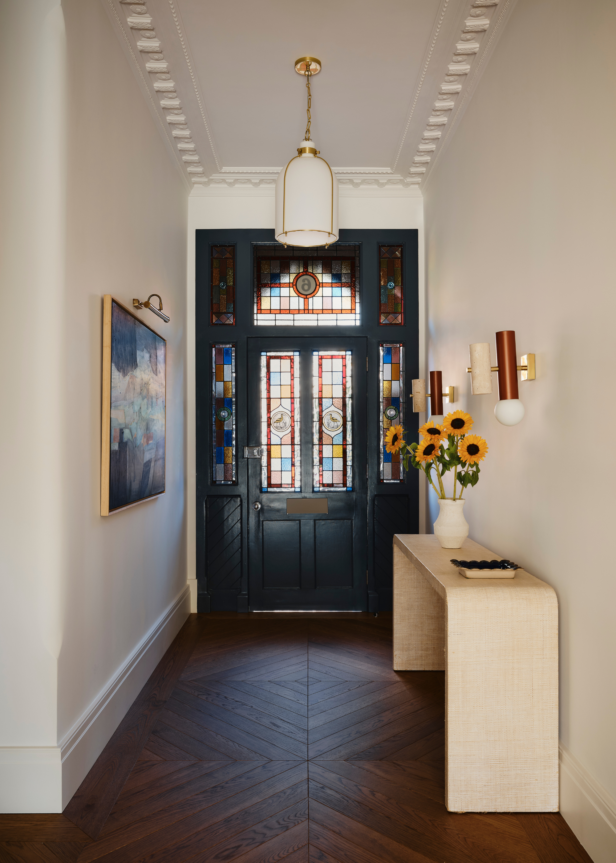 A black painted front door with stained glass panels on and around it with a view of the entryway that has a side table with a vase and sunflowers inside. There is also artwork hanging on the left side of the wall.