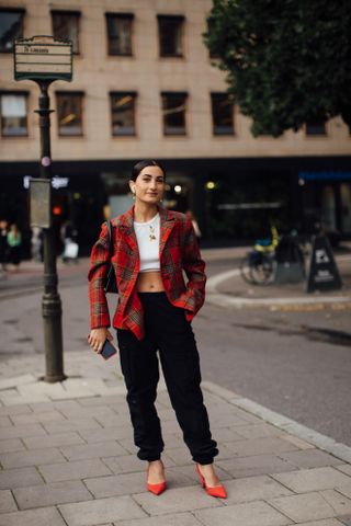 A woman at fashion week wearing a flannel shirt outfit featuring a red plaid blazer, white tee shirt, black pants, and orange heels.