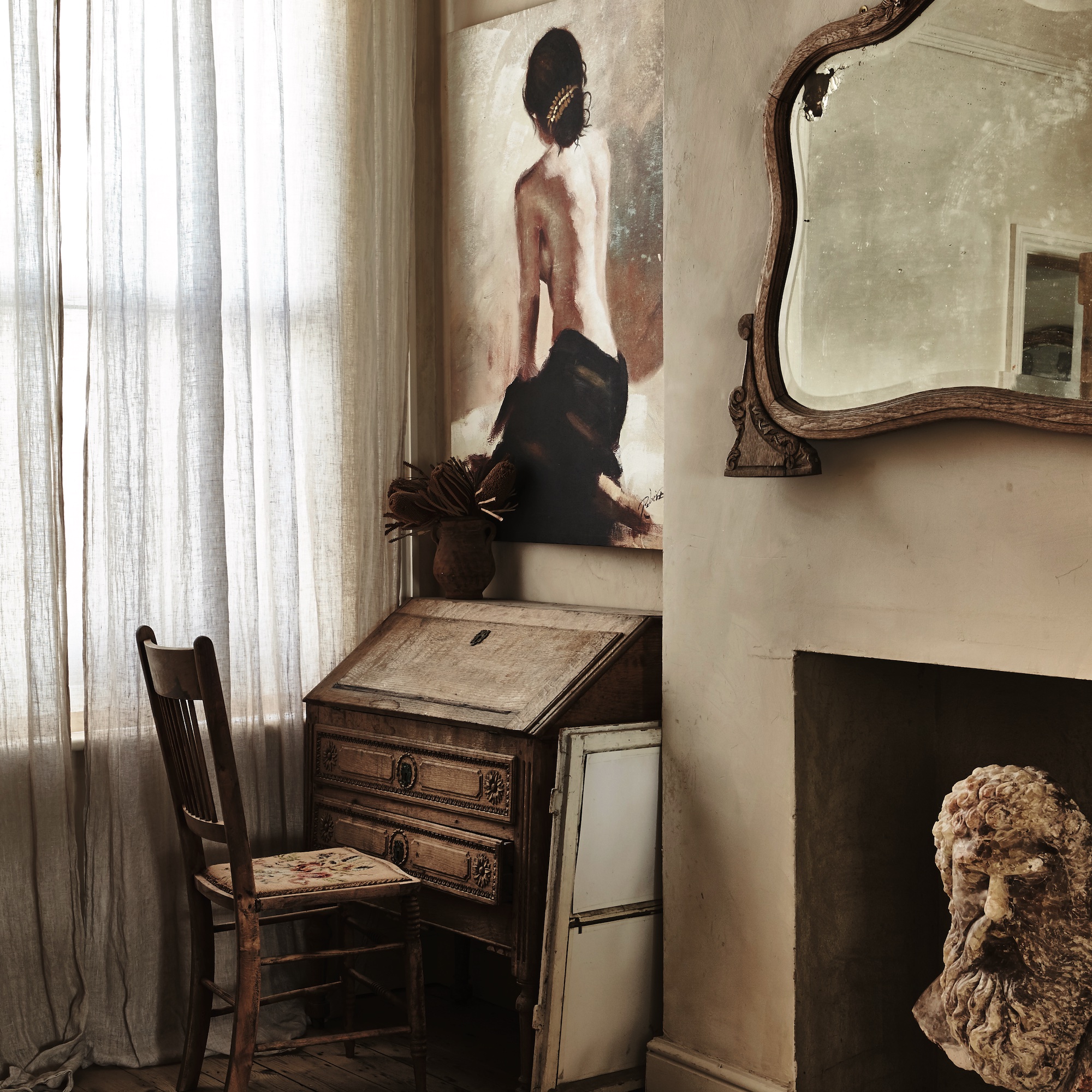 corner of sitting room with vintage wooden bureau in alcove and oil painting above, fireplace with plaster head displayed, and antique mirror on wall