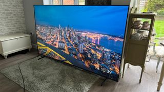 TCL 98Q9BK Mini LED TV in living room on green rug slight angle showing nighttime cityscape on screen