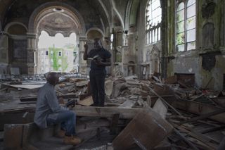 An African man reading a book to a seated African man in a broken down church.