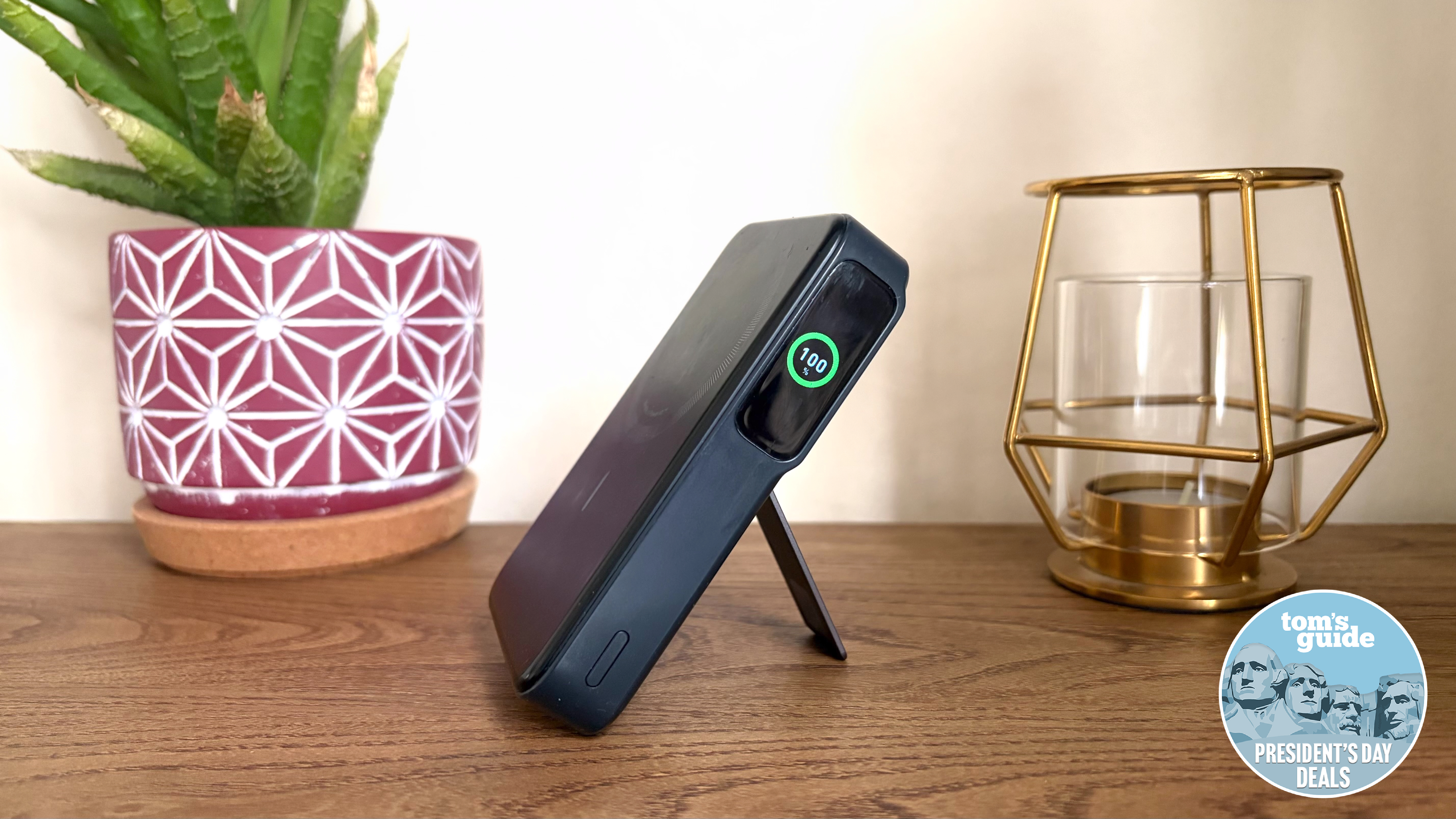 The Anker MagGo Power Bank 10K propped up on its kickstand with the display illuminated showing it has 100% battery on a wooden shelf next to a succulent plant in a red patterned pot and a golden candle holder