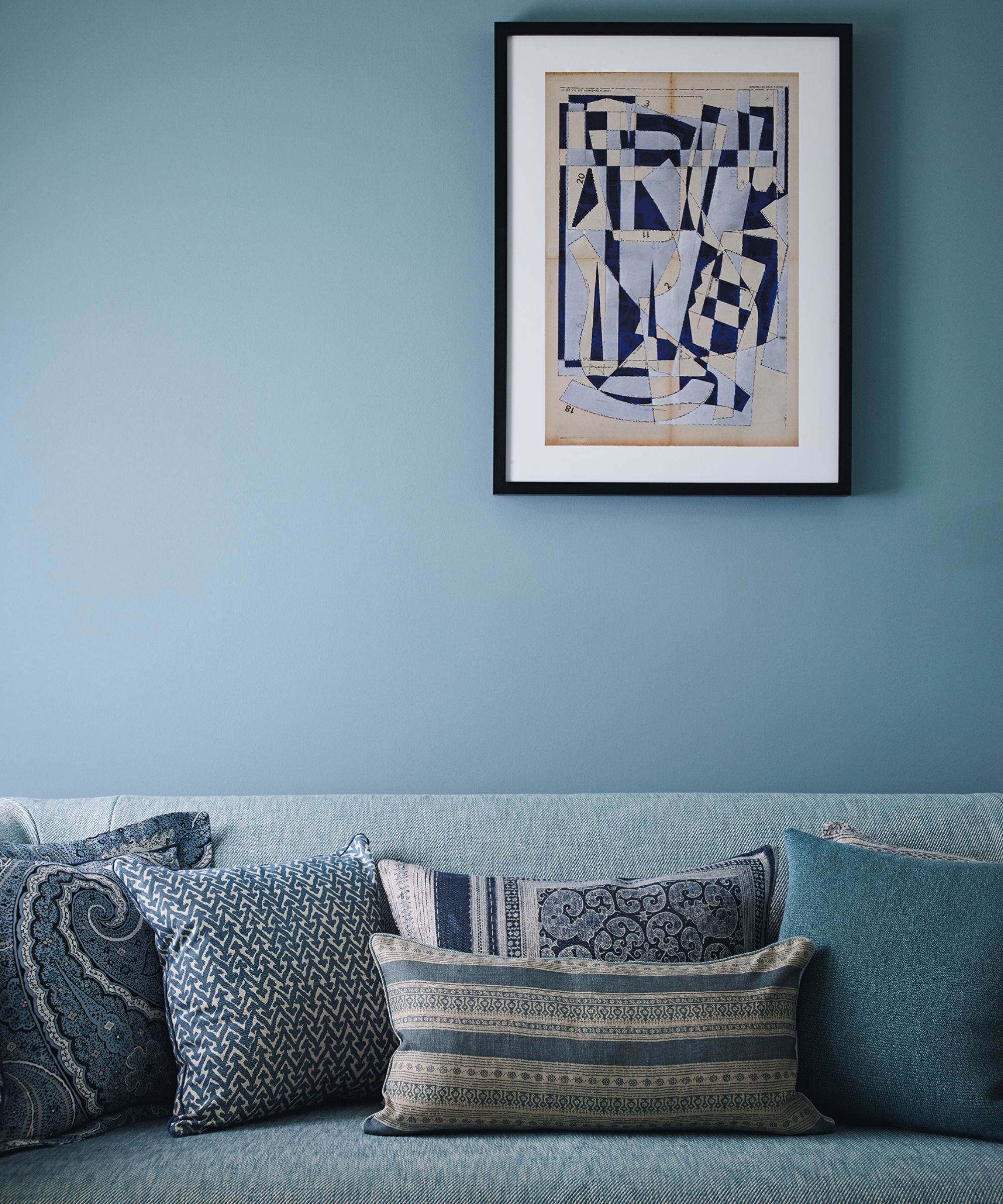 A blue sofa with assorted blue patterned cushions with a blue wall behind and a blue artwork in a black frame above