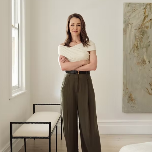 A woman standing with her arms folded wearing dark green trousers and a white top. She is standing in a white room, and there is a white and black bench to the side and an abstract art piece on the wall.