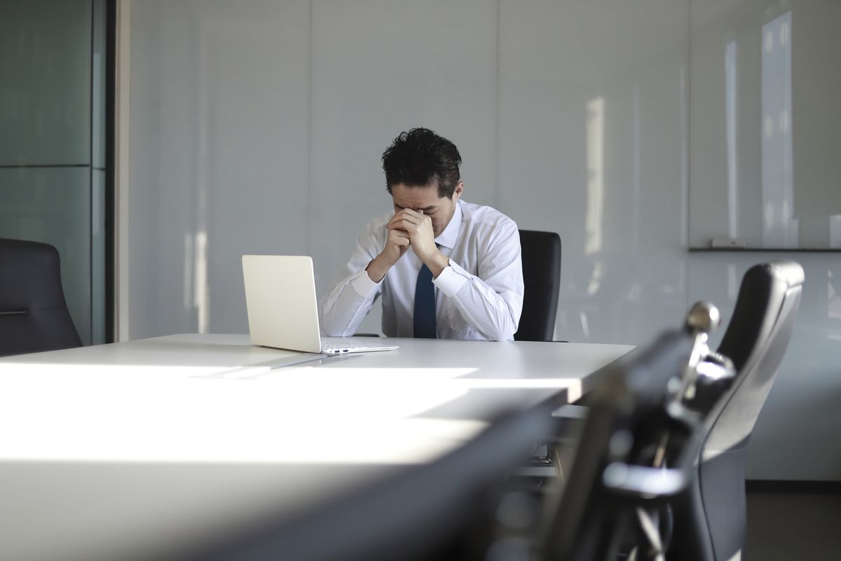 A stressed looking businessman sits at a boardroom table, wearing a suit and tie and holding his head in his hands. A white laptop sits on the table in front of him.