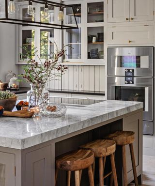 Fitted kitchen with pale grey cupboards and a kitchen island with marbled worktop and wooden stools under the breakfast bar