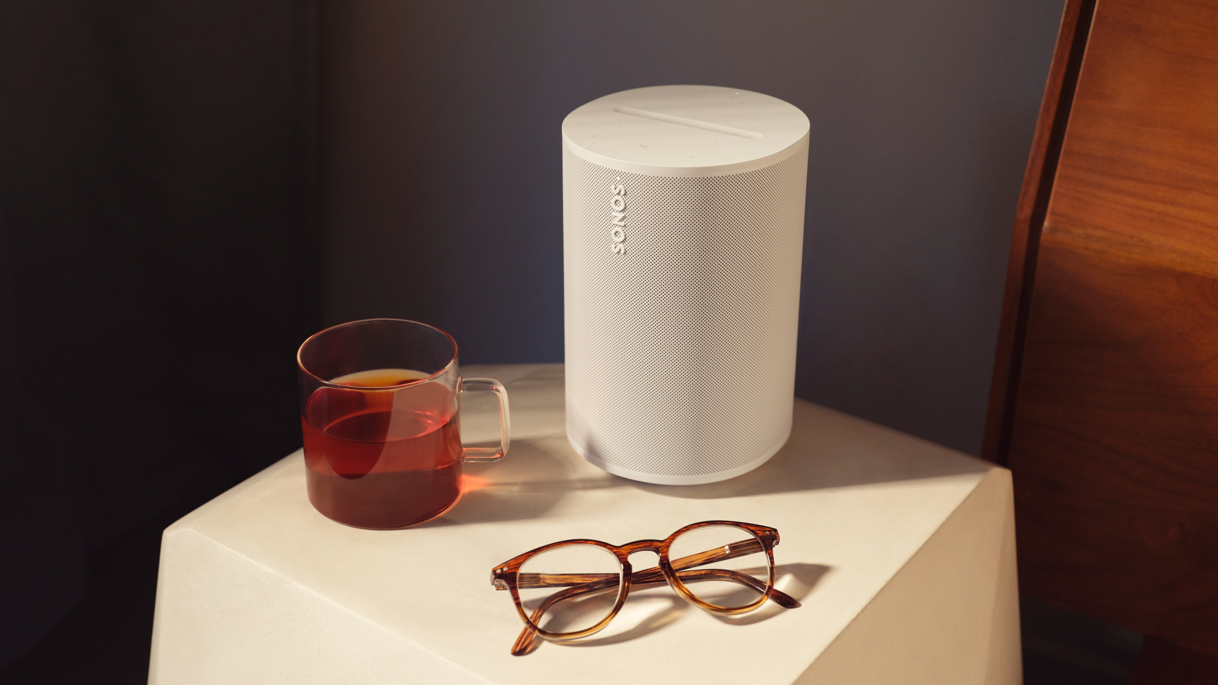 En vit Sonos Era 100 står på ett nattduksbord bredvid ett par glasögon och en tekopp.