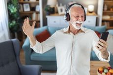Middle aged man joyfully dancing indoors wearing headphones holding a smartphone enjoying music and expressing happiness in a modern home.