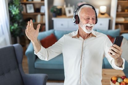 Middle aged man joyfully dancing indoors wearing headphones holding a smartphone enjoying music and expressing happiness in a modern home.