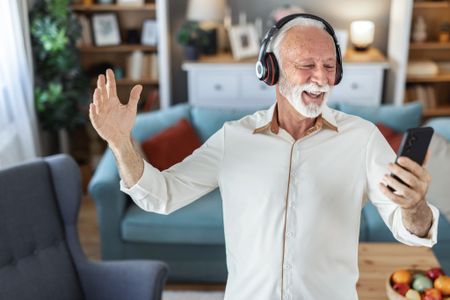 Middle aged man joyfully dancing indoors wearing headphones holding a smartphone enjoying music and expressing happiness in a modern home.