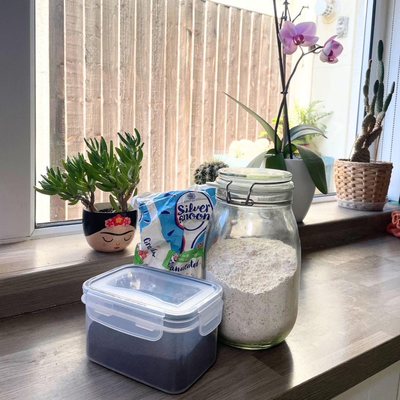 Tupperware of coffee grounds next to a jar of flour and a bag of flour on a kitchen worktop