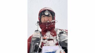 A man in heavy-weather clothing and a hydration vest looks at the camera, covered in snow.