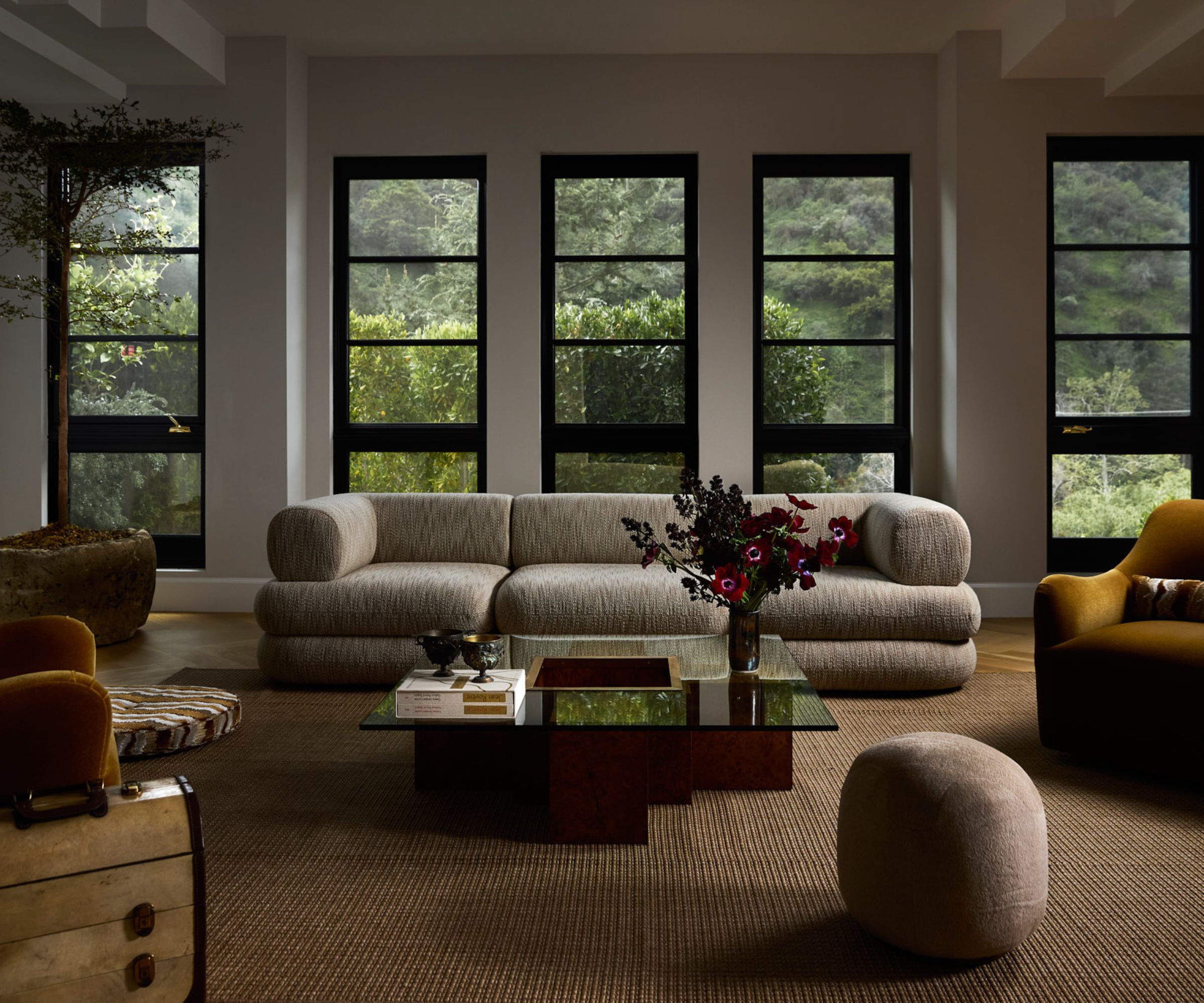 Large modern living room, large cream sofa with yellow armchairs and glass and wood coffee table surrounding, beige carpet and small ottoman, large windows with greenery outside behind