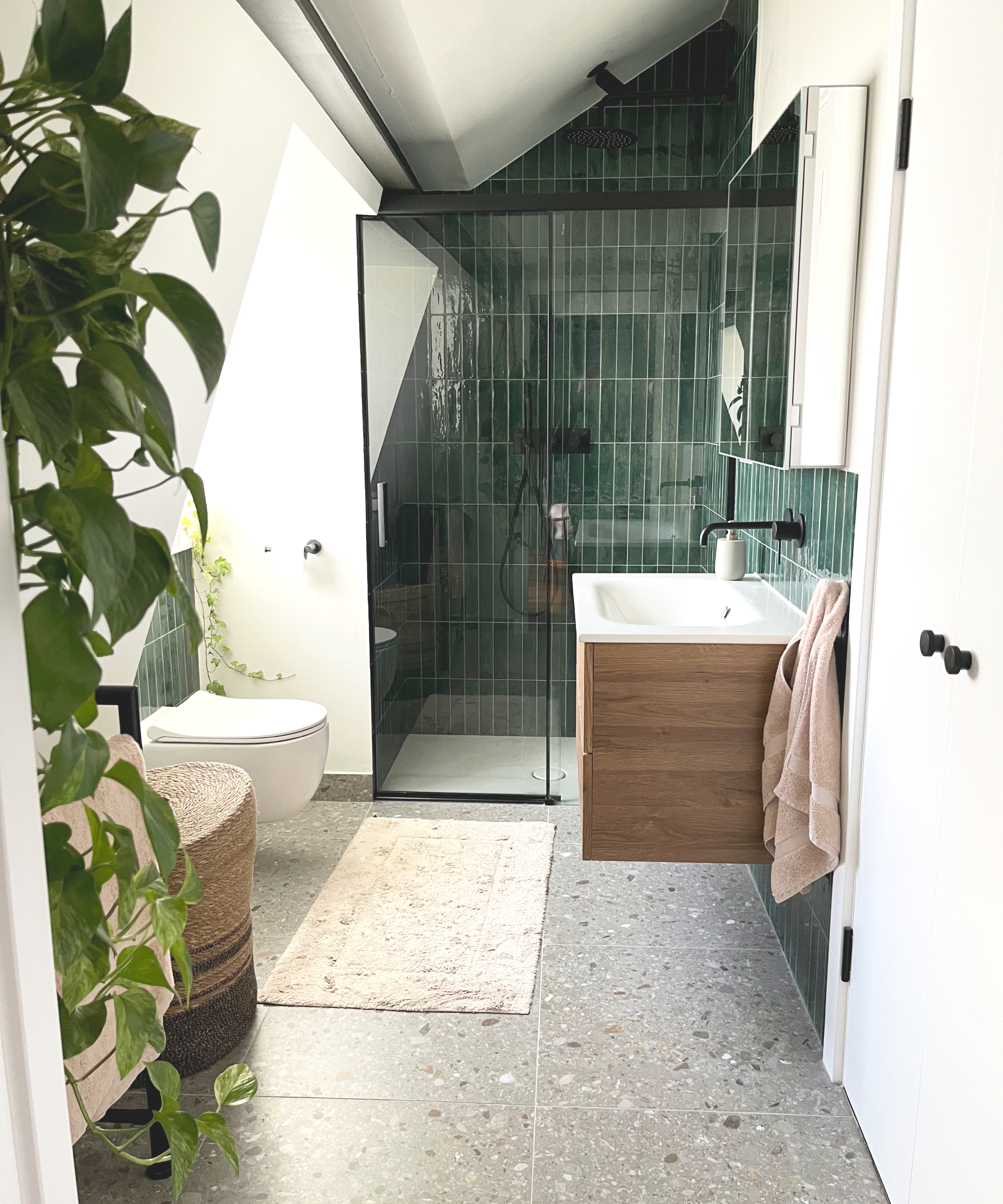 loft conversion bathroom with toilet positioned in space created by the use of a dormer window, dark green tiled walk in shower under the eaves of the roof, a grey terrazzo tiled floor with wall mounted vanity and sink, green plants and mirrored bathroom cabinet