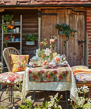 An outdoor dining space with a table decorated with floral table cloth and chairs with colourful cushions