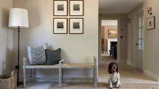 neutral coloured hallway with floor lamp, black and white artwork, bench seat and brown and white spaniel dog sat on hallway runner on top of tiled floor