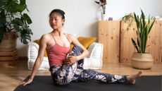 woman with eyes closed facing the camera in a seated twist position wearing floral leggings and pink vest in a living room setting with plants in the corners 