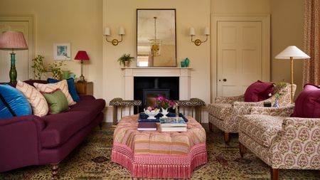 A drawing room in a traditional country home with off-white walls, a fireplace with a wood burner, antiqued mirror with brass wall sconces, a plum sofa, two patterned armchairs, and a pink patterned ottoman.