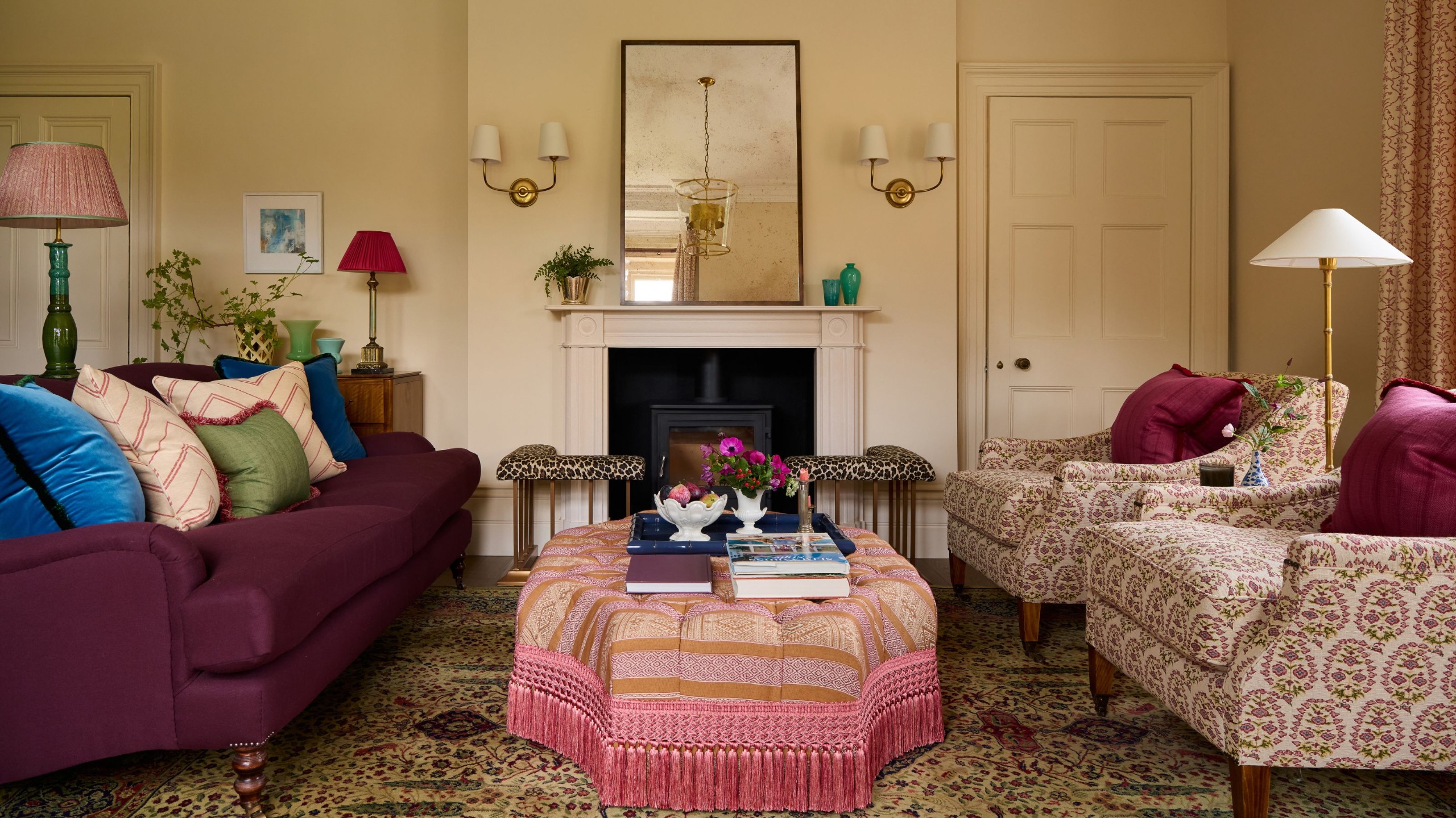 A drawing room in a traditional country home with off-white walls, a fireplace with a wood burner, antiqued mirror with brass wall sconces, a plum sofa, two patterned armchairs, and a pink patterned ottoman.