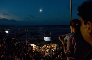 a total solar eclipse unfolds, plunging the ganges river into darkness with people watching on.