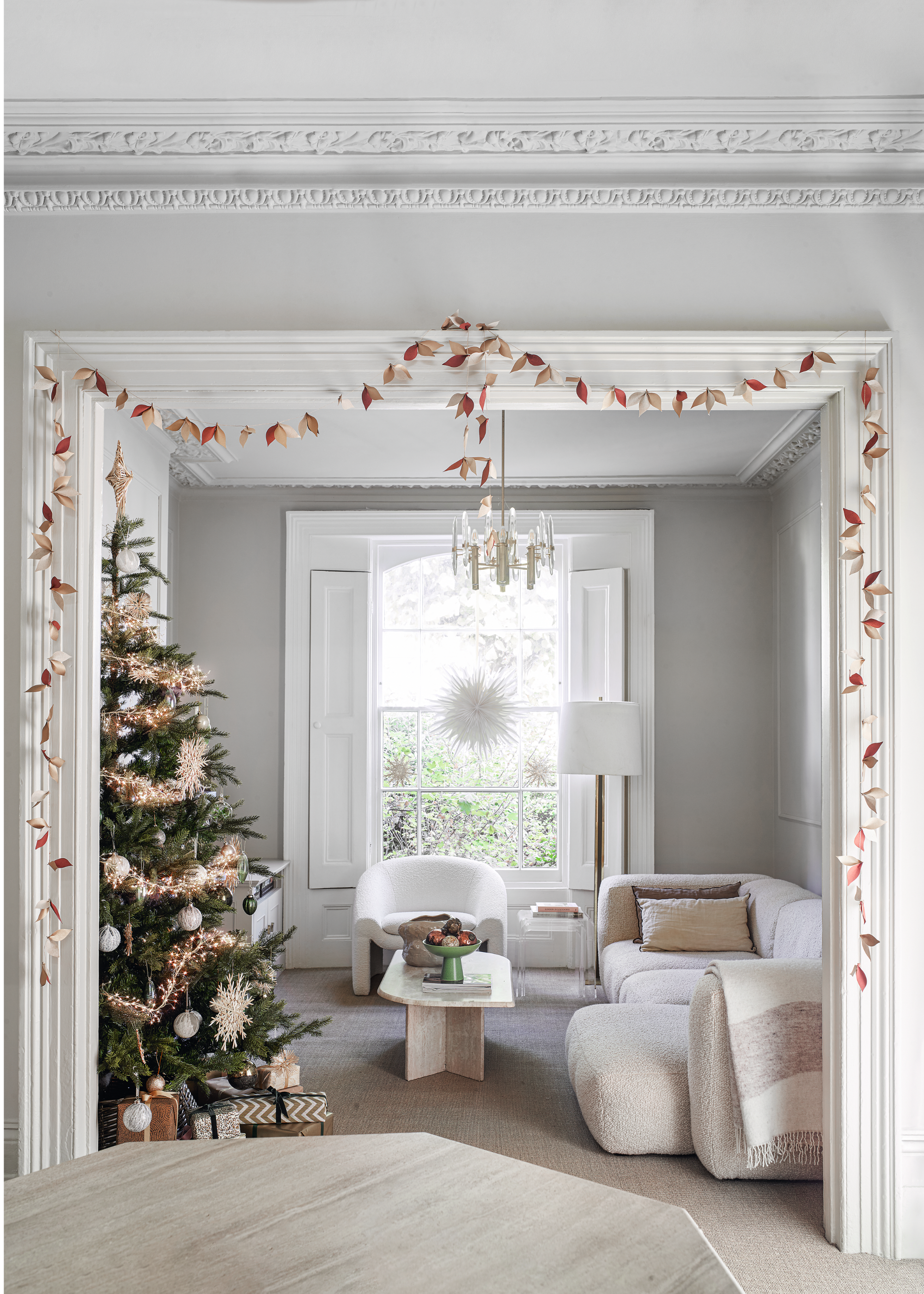A delicate leaf garland is draped across a frame between two living areas in a neutral-decorated space with a christmas tree in the back room deocrated in the beige, cream and orange colors of the garland