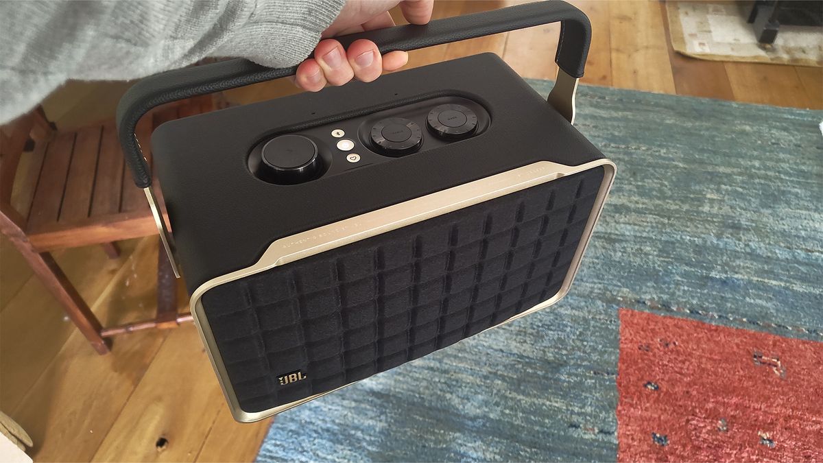 A shot of a black JBL Authentics 300 wireless speaker being held by its integrated handle. A rug can be seen on the floor in the background.