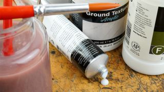 A tube of acrylic gloss medium with other tubs of medium and a jar of water