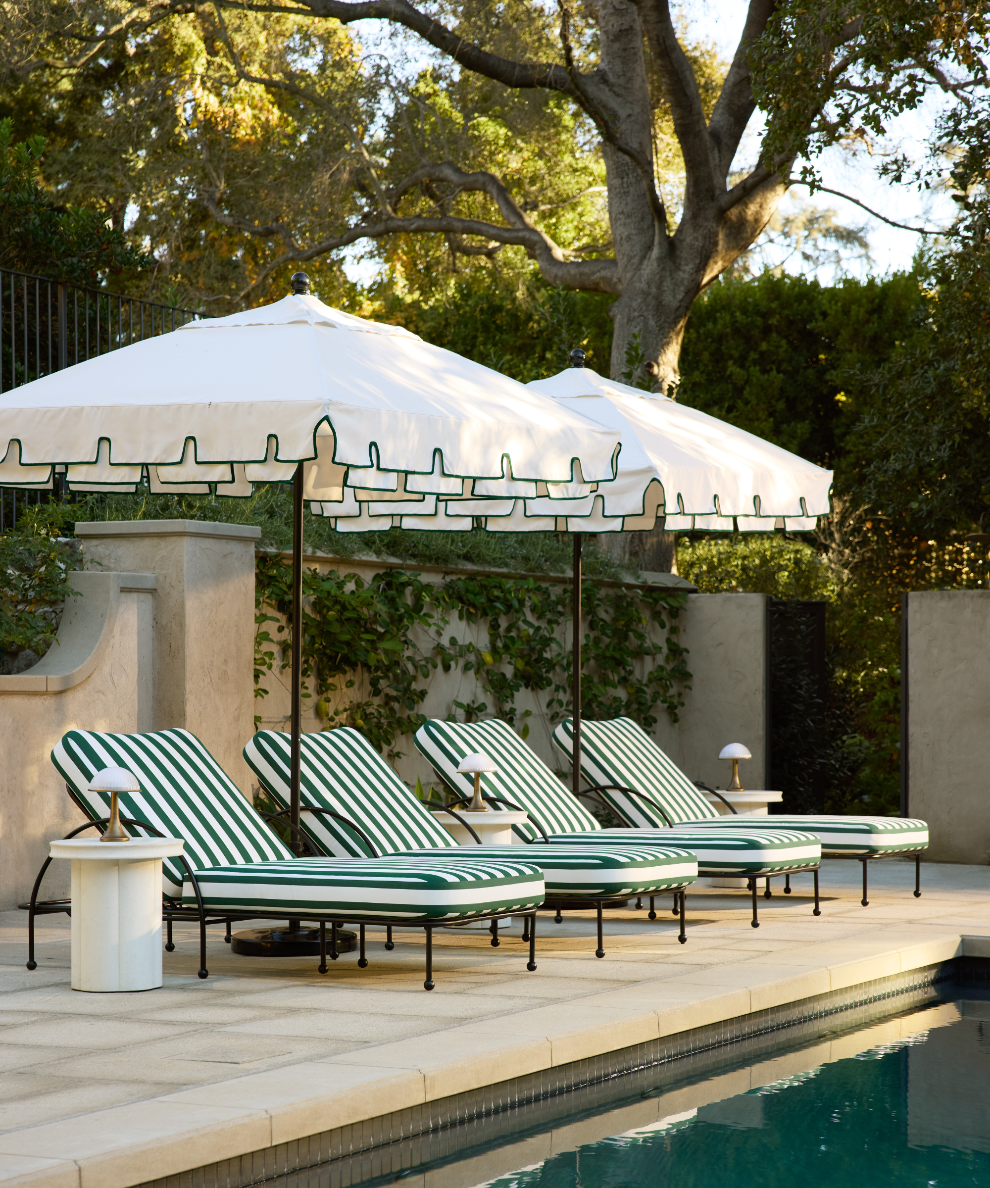 a poolside set up with four lounge chairs with striped cushions, two umbrellas with green trip, and side tables with lamps on each one