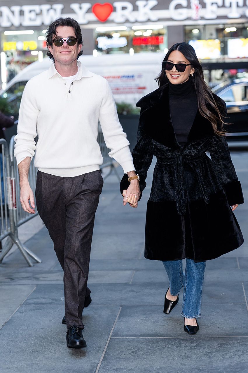 Olivia Munn with John Mulaney, wearing a black coat and turtleneck, stovepipe blue jeans, and black pumps