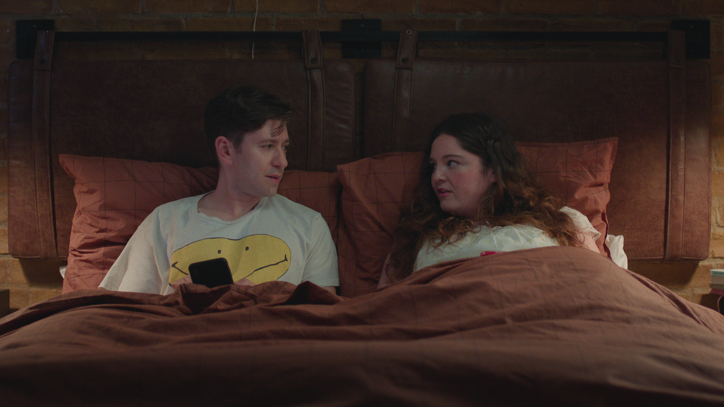 michael zegan as zev and meg stalter as jessica lying in bed together in the tv show too much