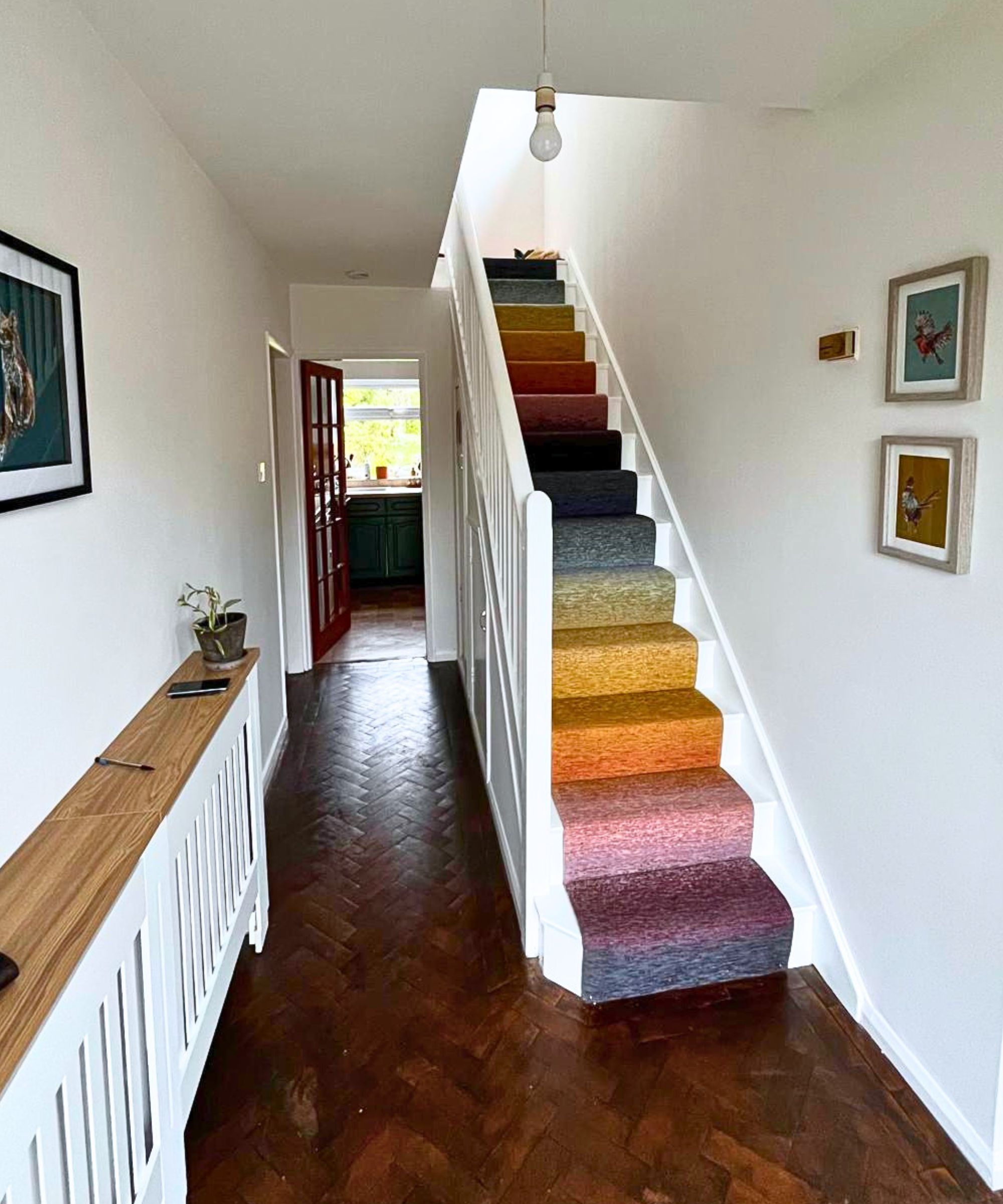 Dark herringbone parquet floor, white walls with framed art, and a striking rainbow ombr&eacute; stair carpet graduating from purple at the bottom through orange, yellow and grey to dark at the top.