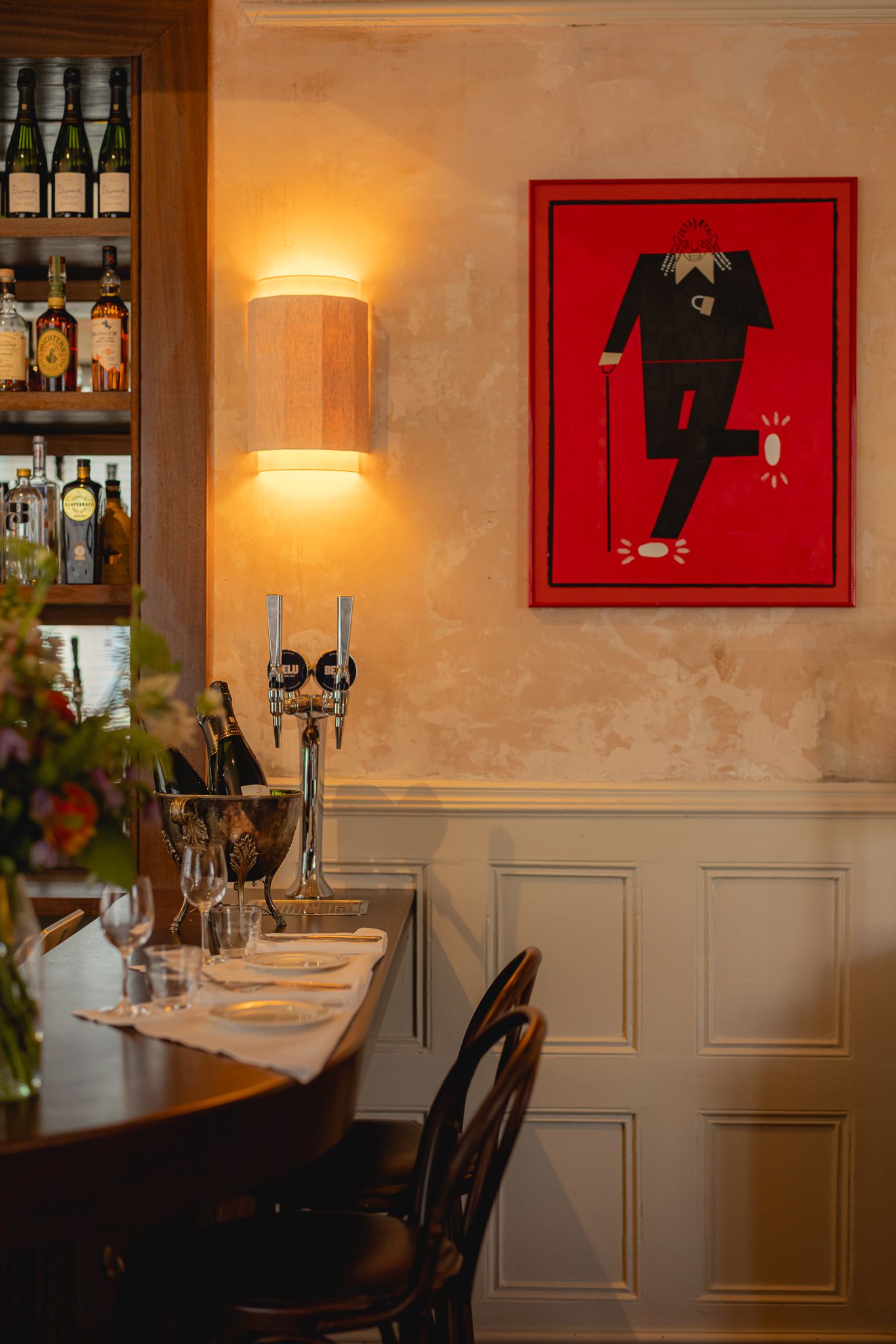 The charming-cool interiors of a French-style modern bistro with distressed plaster walls, lived-in wooden floors, elegant tables and chairs, colorful artworks, beige and red curtains, and sunset-hued sconces and pendant lights.