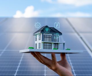 hand holding model of house with energy icons around it with solar panels behind