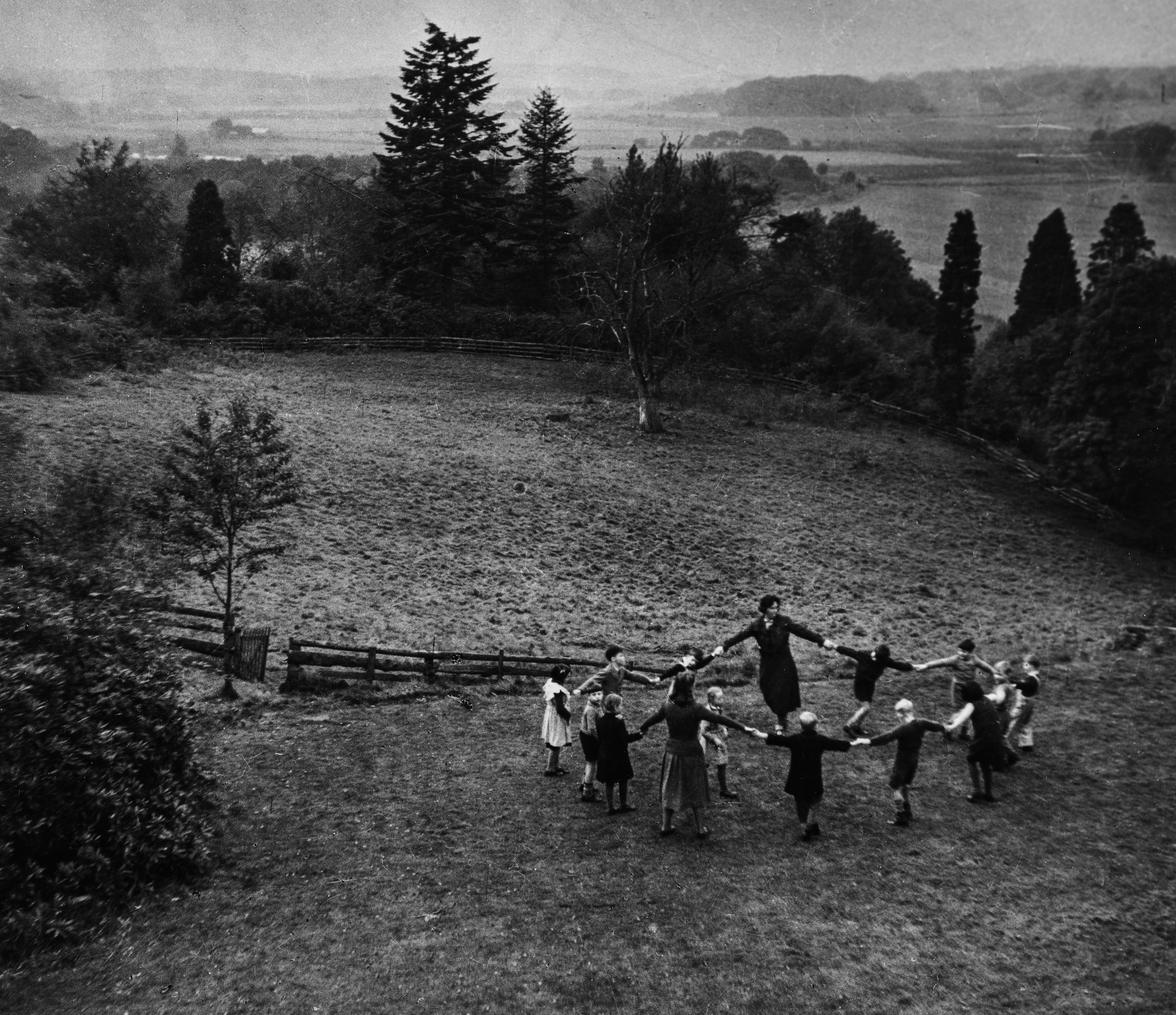 Viewed from a high vantage point, a circle of children and one adult hold hands in a ring on a wide expanse of open grassland. Tall conifers and rolling countryside stretch into the misty distance. The figures are small against the landscape, giving the image a lyrical, almost pastoral quality.