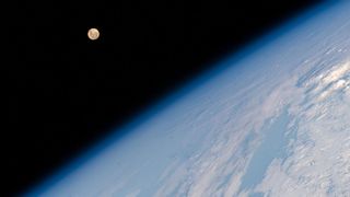 the curvature of a rocky blue earth fills the bottom left to top right of the photo at a diagonal. A small full moon hangs in the upper left, just to the right, in a black abyss.