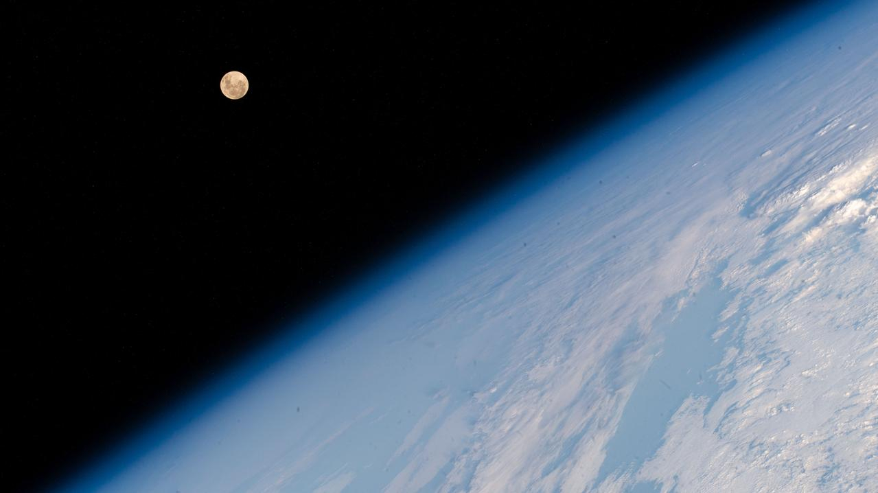 the curvature of a rocky blue earth fills the bottom left to top right of the photo at a diagonal. A small full moon hangs in the upper left, just to the right, in a black abyss.