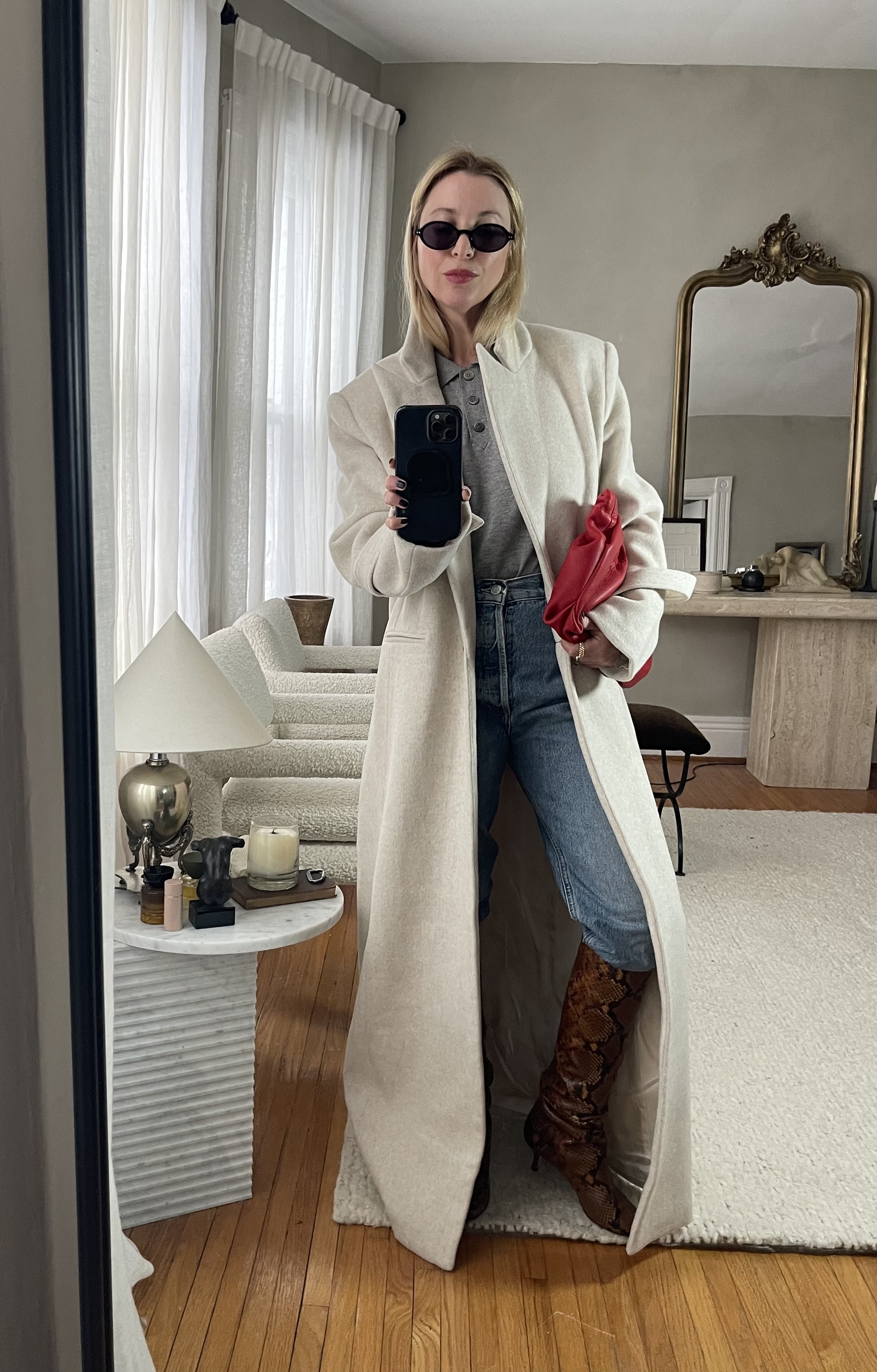 Kerry Pieri wearing a white coat, grey polo sweater, blue jeans, brown boots, and a red clutch