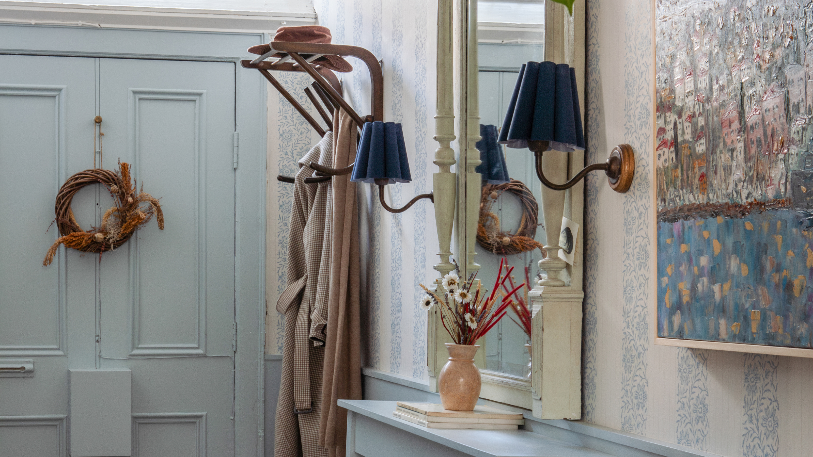 a small entryway with a blue painted door and a wreath hanging on it, a small coat hook mounted to the wall, and two sconces on either side of a full length mirror 