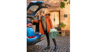 A blonde-haired woman wearing a coat and holding the Mamas and Papas 3-in-1 Floor & Booster Seat with Activity Tray in her left hand stands by a blue car with the boot open, loading luggage into the vehicle