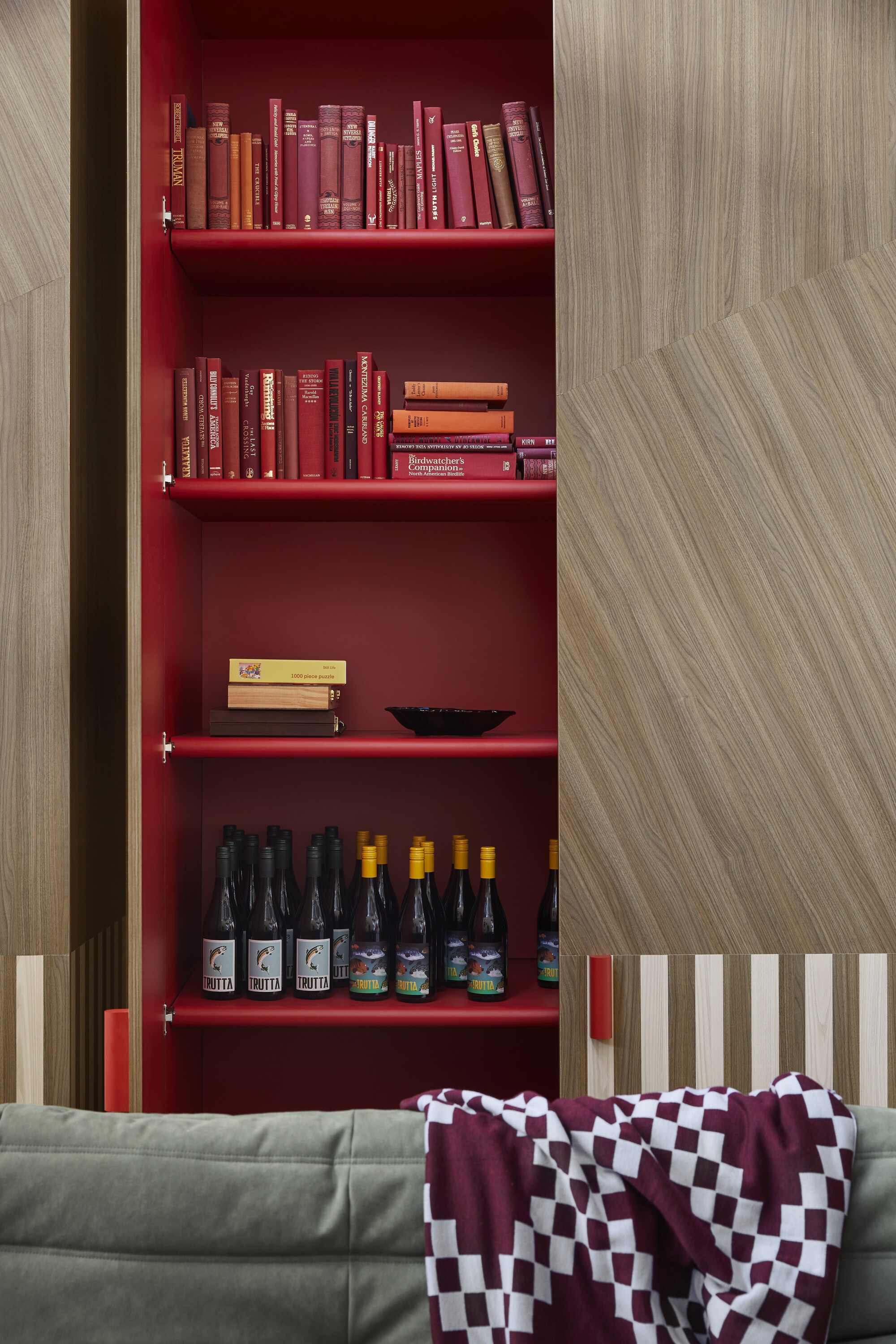 Image of a wooden closet with a sliding door that is open to reveal a red interior. The shelves of the closet are filled with red books and wine bottles. There is the top of an army green sofa in view with a purple and white checkered throw draped on it.