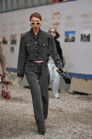 A guest wears white latte vintage sunglasses, black and crystals earrings, a dark gray denim with embroidered crystals buttoned jacket, matching dark gray denim with embroidered crystals wide legs pants, a black nylon nailed / studded handbag, silver shiny varnished leather pointed pumps heels shoes , outside Munthe, during the Copenhagen Fashion Week Spring/Summer 2024 on August 10, 2023 in Copenhagen, Denmark.