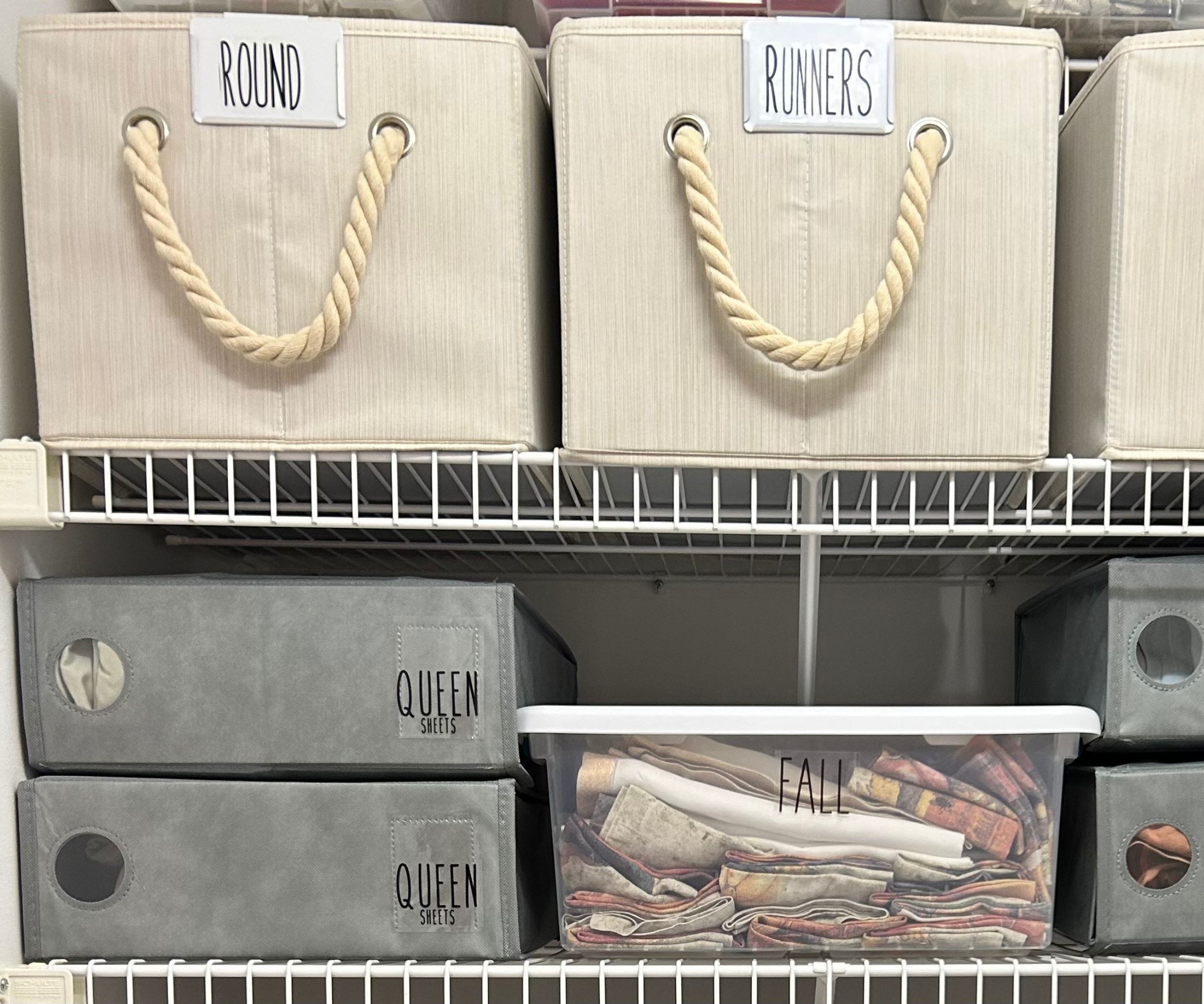 Grey bedding storage boxes on wite shelving, underneath cream labelled baskets with rope handles.