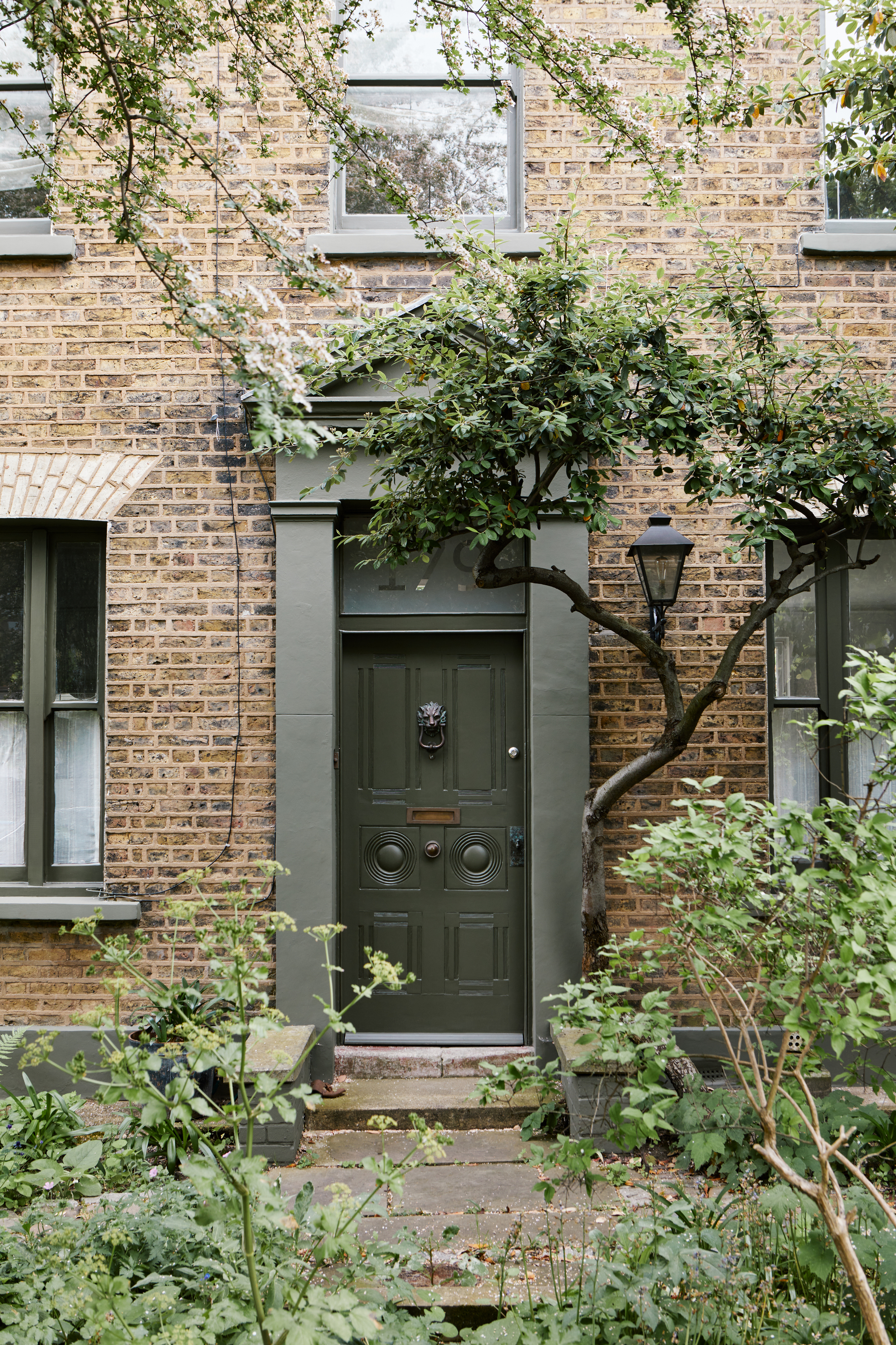 Image of a dark green front door on a light brick home. The window trims and trim around the door are all sage green. There are lots of plants and trees around the house.