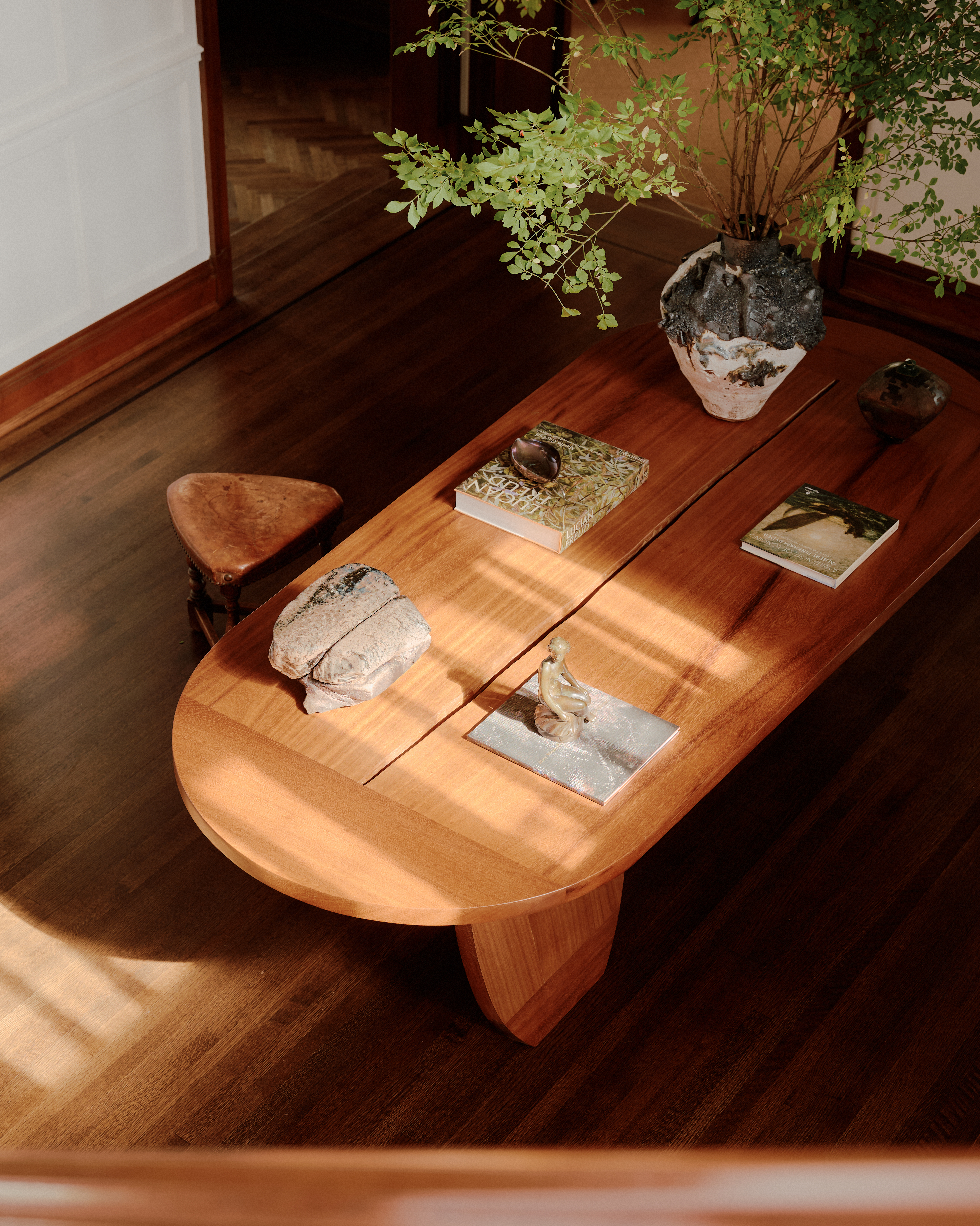 An oblong wooden table made by hand features a central crack, chubby legs, and lots of ceramic sculptures placed on it as it bathes in sunshine.