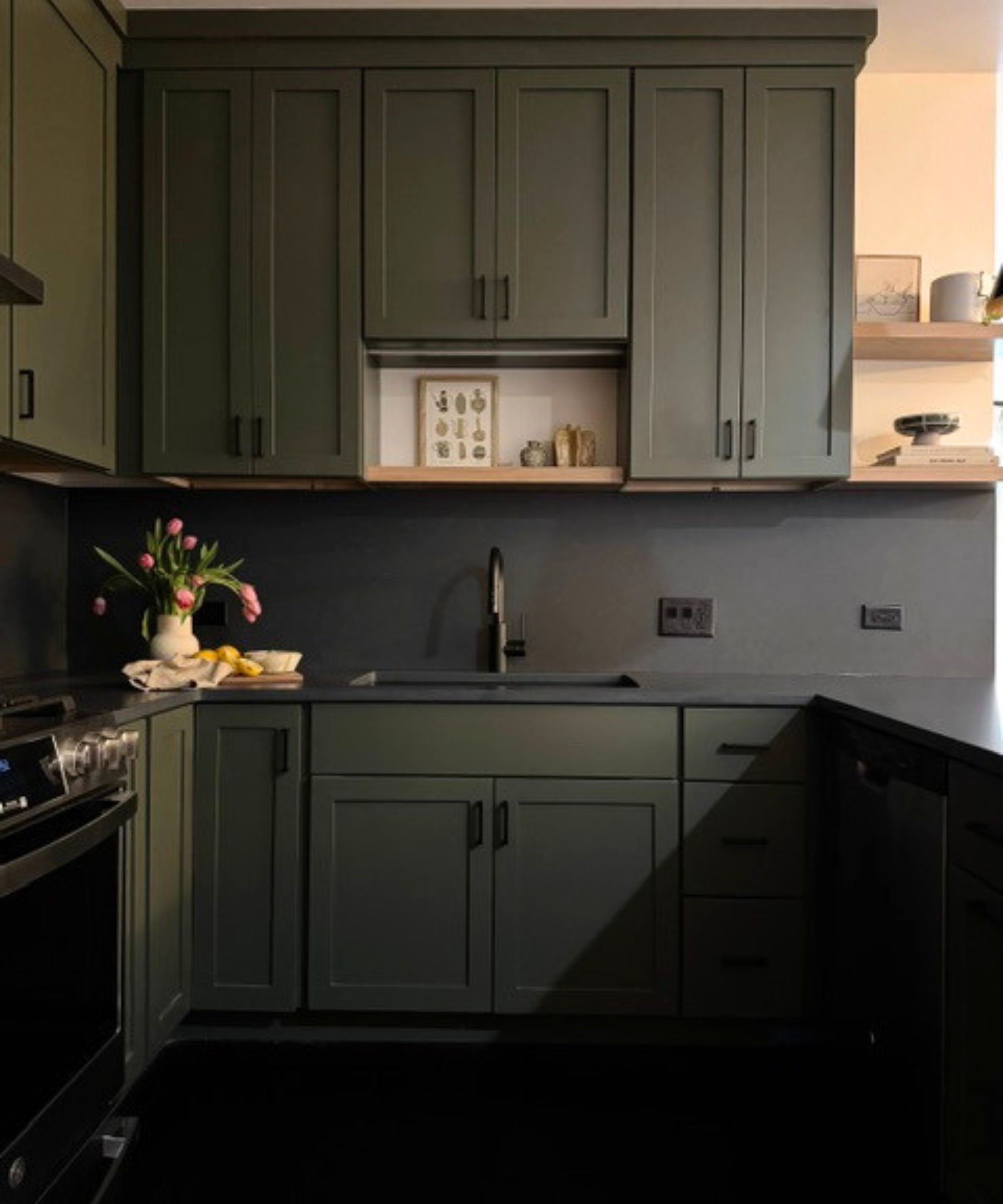 Kitchen with Pewter Green by Sherwin-Williams painted cabinetry, a black backsplash and countertops, a range oven, wooden open shelves with ceramics and prints, and a vase of tulips.