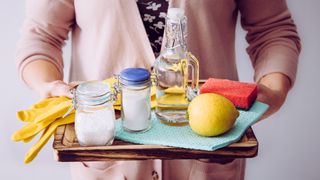 Woman holding tray with washing up gloves, sponge, cloth, lemons and vinegar to use for how to clean windows with natural products