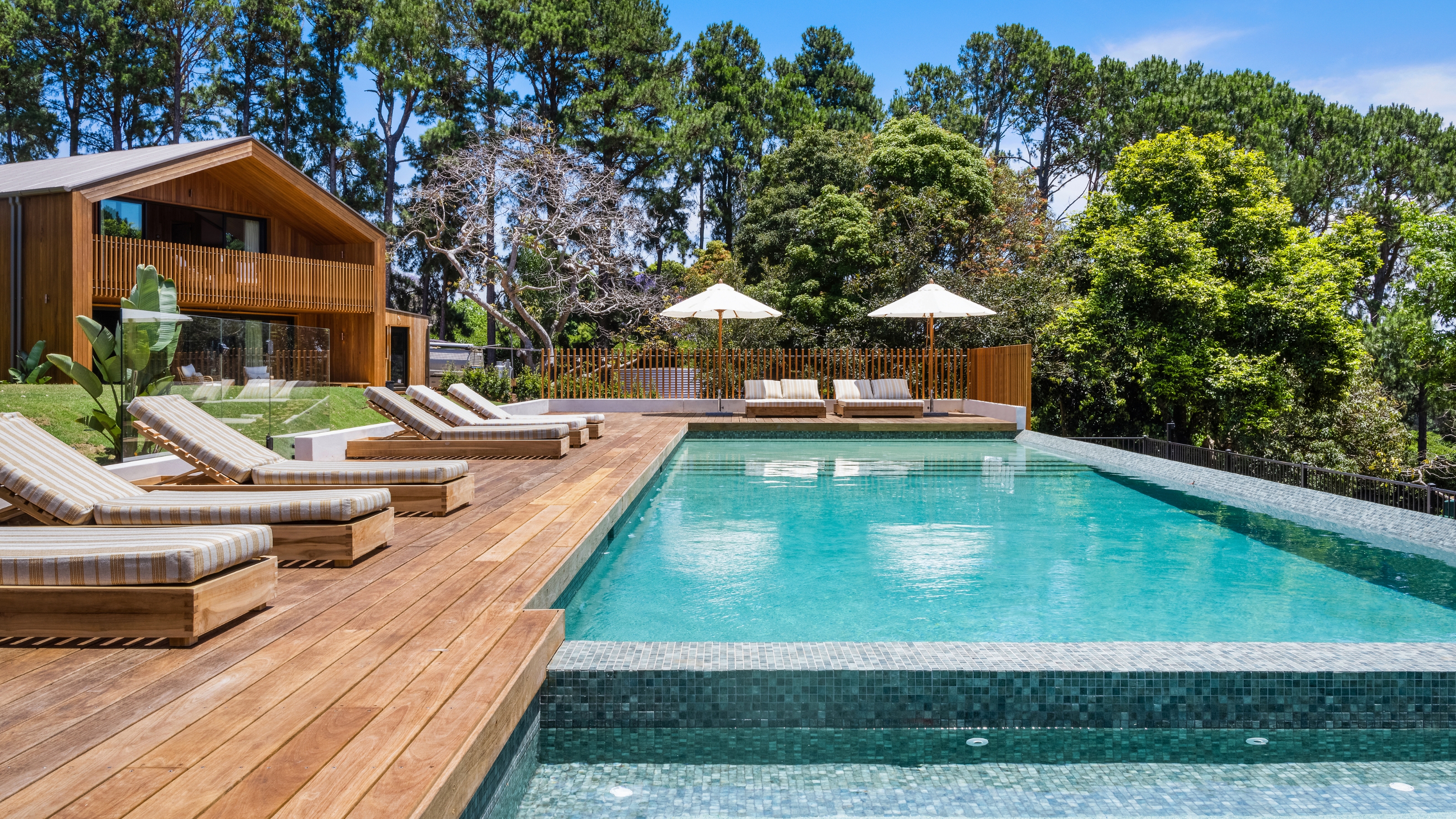 Image of a pool and pool deck in the direct sunlight with multiple lounge chairs and two umbrellas.